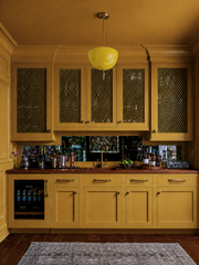 See the handmade yellow Alexander Pendant styled in a moody mustard bar kitchenette — its glossy powder-coated dome and brass finial glowing like a drop of sunshine against tonal ochre cabinetry, brass mesh cabinet fronts, and an antiqued mirror backsplash. This is color-drenching done right: a bright yellow light fixture layered into a warm, saturated room where wood counters and aged brass keep everything grounded. Perfect inspiration for a butler's pantry, home bar, or maximalist kitchen. Shop handmade y