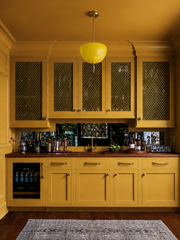 Few rooms commit like this one: mustard yellow poured over the walls, ceiling, and cabinetry for an enveloping, tonal effect that feels warm and a little vintage. Brass mesh cabinet fronts and an antiqued mirror backsplash add shine and old-world texture, while walnut counters and a well-stocked bar keep it grounded and livable. The yellow Alexander Pendant is the exclamation point, its glossy domed shade glowing like a drop of sunshine against the deeper gold tones. A bold, saturated look for anyone who lo