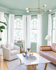 Sage green walls frame tall arched windows dressed in white linen curtains. A brass chandelier with opal glass shades hangs centrally. A cognac leather sofa sits opposite a cream upholstered chair, with a marble-topped brass table between them holding stacked books and fresh flowers. Hardwood floors, potted plant on left, floor lamp on right with beige shade.
