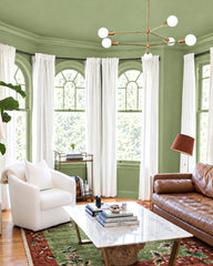 Sage green walls with tall arched windows and white linen curtains. A white opal globe pendant hangs from brass hardware. A patterned rug in deep green and rust botanical print covers the floor. Cognac leather sofa and cream upholstered chair positioned across from each other. Marble-topped brass table between them with stacked books and flowers in a clear vase.