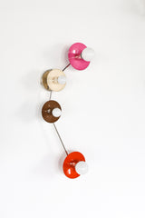 Colorblocked Aries wall sconce with four round saucer disc shades in doll pink, cream, fudge brown, and flame orange, connected by sleek chrome arms with white globe bulbs, photographed on a clean white background. Sculptural modern lighting for a pretty powder room, feminine bathroom vanity, eclectic bedroom, colorful hallway, mid century modern living room, or playful kids room, an instant pop of color for maximalist and modern decor.