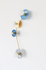 Colorblocked Aries wall sconce with four round saucer disc shades in hydrangea blue, white, denim blue, and slate grey, paired with warm brass arms and white globe bulbs on a clean white background. Sculptural modern lighting with a mid century modern vibe for a bathroom vanity, pretty powder room, hallway, bedroom, entryway, or kids room. Great for coastal decor, Scandinavian style interiors, eclectic color-forward homes, and designer lighting projects.