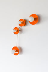 Angled view of Aries wall sconce in Orange Peel, a high energy glossy orange modern wall light with chrome arms and chrome canopy, four round disc shades with exposed white globe bulbs, sculptural geometric sconce that reads like wall art, perfect statement lighting for a modern entryway, eclectic hallway, colorful living room, or maximalist bedroom, mid century modern style wall fixture with playful proportions, bright orange accent lighting for dopamine decor and colorful interior design, contemporary art