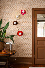 Colorblocked Aries wall sconce with four saucer disc shades in doll pink, cream, fudge brown, and flame orange, connected by sleek chrome arms with white globe bulbs, shown as a plug in wall light with cord. Styled on warm tan floral wallpaper next to a brown paneled door with glass, a vintage wood side table, a chrome table lamp, and a large leafy houseplant. Retro eclectic decor for a cozy entryway, hallway, stair landing, reading nook, or moody powder room, perfect for maximalist and mid century modern
