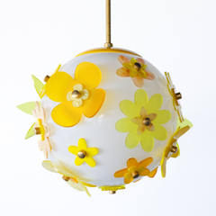 One-of-a-kind floral acrylic globe pendant light on white background, white satin-diffuse sphere wrapped in layered yellow and chartreuse acrylic flowers with warm metal caps, suspended from a warm brass stem; cheerful sculptural statement made in New Orleans.