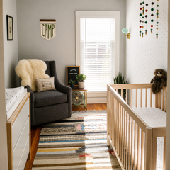 Modern baby boy nursery with a mint green Loa wall sconce in brass over a light wood crib and patterned accent wall. Warm, neutral styling includes a cozy grey glider, striped rug, plants, and playful decor accents. Great for nursery lighting ideas, mint wall sconce, brass sconce, modern nursery design, and gender-neutral baby room decor.