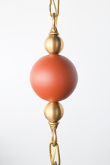 Closeup of powdercoated terra cotta ball on beaded chain on a One of a kind cream pendant light with a sculptural hourglass shade hanging from a brass beaded chain, featuring terracotta round beads and light teakwood accent beads, a modern mid century inspired statement pendant for maximalist design lovers, perfect over a kitchen island, breakfast nook, entryway, nursery, or kids room lighting.