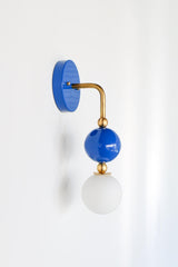 The Ida Sconce pairs a brass hooked arm with a glossy Mediterranean blue glass ball and a soft white opal glass globe at the bulb, giving it a sculptural, almost jewelry-like presence on the wall. It works well as a bedside light in place of a table lamp, beside a bathroom mirror, or anywhere a single statement sconce makes more sense than a matching pair.
