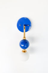 The Ida Sconce pairs a brass hooked arm with a glossy Mediterranean blue glass ball and a soft white opal glass globe at the bulb, giving it a sculptural, almost jewelry-like presence on the wall. It works well as a bedside light in place of a table lamp, beside a bathroom mirror, or anywhere a single statement sconce makes more sense than a matching pair.
