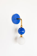 The Ida Sconce pairs a brass hooked arm with a glossy Mediterranean blue glass ball and a soft white opal glass globe at the bulb, giving it a sculptural, almost jewelry-like presence on the wall. It works well as a bedside light in place of a table lamp, beside a bathroom mirror, or anywhere a single statement sconce makes more sense than a matching pair.