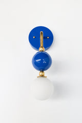 The Ida Sconce pairs a brass hooked arm with a glossy Mediterranean blue glass ball and a soft white opal glass globe at the bulb, giving it a sculptural, almost jewelry-like presence on the wall. It works well as a bedside light in place of a table lamp, beside a bathroom mirror, or anywhere a single statement sconce makes more sense than a matching pair.