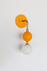The Beaded Sconce in mustard stacks a hooked brass arm, small brass ball joints, and a matte mustard glass sphere above a soft white opal glass globe, giving the whole fixture a jewelry-like, beaded silhouette on the wall. It works well as a bedside light in place of a table lamp, beside a bathroom mirror, or anywhere a single statement sconce makes more sense than a matching pair.