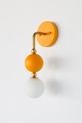 The Beaded Sconce in mustard stacks a hooked brass arm, small brass ball joints, and a matte mustard glass sphere above a soft white opal glass globe, giving the whole fixture a jewelry-like, beaded silhouette on the wall. It works well as a bedside light in place of a table lamp, beside a bathroom mirror, or anywhere a single statement sconce makes more sense than a matching pair.