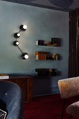 Moody interior photo of the Aries wall sconce in an all-black finish, a sculptural modern wall light with four round disc shades and white globe bulbs arranged along an angled arm like graphic wall art. Mounted on a deep, smoky blue textured wall (plaster/limewash look) with warm light glow and soft shadows. Styled in an eclectic, masculine reading room or home office with floating shelves holding vintage books and a dark wood cabinet. Statement black wall lighting for a hallway, entryway, den, media room, 