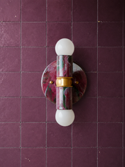 A two-light marbled sconce in black cherry, olive, and lilac against deep aubergine square tile with a slightly worn, old-house glaze. Brass hardware joins the upper and lower arms, each holding an exposed globe bulb. A useful pairing reference for anyone building a moody, colorful bathroom instead of a safe neutral one.
