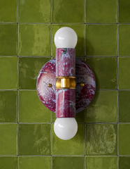 A two-light marbled sconce in black cherry, olive, and lilac set against glossy olive green square tile. Brass hardware connects the upper and lower arms, each with an exposed globe bulb. A strong color-pairing reference for a bathroom or powder room leaning into saturated, unexpected combinations instead of playing it safe.
