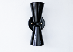 A statement wall sconce for rooms that lead with sculptural, graphic shapes. Seen from an angle, the Dryades sconce shows off its glossy black double-cone silhouette — one cone throwing light up, one down — meeting at a brass-collared waist on a round black backplate. The three-quarter view highlights how it projects off the wall and its crisp mid-century lines. A bold, moody pick for hallways, bars, bedrooms, and Art Deco or masculine interiors.