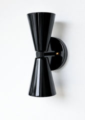 A sculptural double-cone wall sconce for a hallway, bar, bedroom, or mid-century modern room that wants crisp, two-directional light. The Dryades sconce pairs an upward and a downward glossy black cone at a narrow brass-collared waist, mounted on a round black backplate with brass screw accents. Graphic, bold, and a little Art Deco, its bowtie silhouette makes a confident statement in moody, masculine, or modern eclectic spaces.