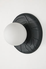 Angled view of matte black unglazed porcelain wall sconce with frosted glass globe, showing sculptural leafy relief texture