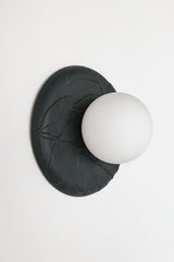 Angled view of matte black unglazed porcelain wall sconce with frosted glass globe, showing sculptural leafy relief texture