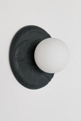 Angled view of matte black unglazed porcelain wall sconce with frosted glass globe, showing sculptural leafy relief texture