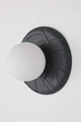 Side view of matte black unglazed porcelain wall sconce with raised leafy relief and frosted glass globe, sculptural modern ceramic lighting