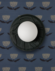 Top-down product photo of the Lierre ceramic globe wall sconce in unglazed matte black with raised leafy relief, centered on blue-and-white woodblock-style floral wallpaper, a moody modern accent light that pairs with traditional pattern, grandmillennial decor, and contemporary contrast.