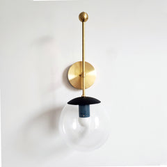 A sculptural clear-globe sconce for a powder room, bedside, or hallway that wants a wall light with a genuinely custom feel. This Verbena Sconce hangs its clear glass globe below a black dome shade, with a navy socket sleeve inside for a subtle two-tone pairing, so the bulb glows downward through the glass. A slim brass stem rises to a small ball finial above the round backplate, a quiet flourish that sets it apart. The black-and-navy combination reads moody and graphic at once, the kind of considered detai