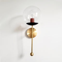 An earthy-meets-graphic clear-globe sconce for a powder room, bedside, or hallway that wants a wall light with a genuinely custom feel. This Verbena Sconce swaps the usual opaque shade for a clear glass globe, so the bulb shows through, seated in a black cup with a warm terracotta socket sleeve inside for an unexpected two-tone pairing. A slim brass stem, round backplate, and small brass ball finish it below. The black-and-terracotta combination reads grounded and a little bold at once, the kind of consider