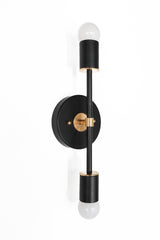 Vertical Medium Thalia wall sconce in matte black with warm brass hardware, featuring a round black backplate, slim center bar, and two exposed frosted globe bulbs top and bottom with brass accents, modern black and brass wall light for bathroom vanity sconces, entryway and hallway lighting, bedroom bedside wall sconces, and design forward contemporary or mid century modern home decor