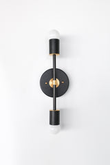 Front view of the Medium Thalia Sconce in matte black with warm brass hardware, a modern vertical double wall sconce with two exposed globe bulbs and a round backplate, sleek mid century modern bathroom vanity light or bedside wall light for contemporary, eclectic, and design-forward interiors.