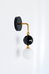 A single-arm wall sconce with a glossy black round backplate and a brass gooseneck arm that curves down to a black glass orb, small brass ball accents, and a frosted globe bulb below. The stacked, sculptural shape gives it more graphic presence than a standard sconce, a strong pick for a moody bathroom, a hallway, or anywhere that wants one confident lighting moment.