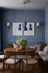 Moody blue dining room with paneled walls, built-in banquette seating, mid century wood dining chairs, abstract line art, striped pillows, and a round wood table. The Blue Margot Chandelier hangs from a brass chain above the table, adding a sculptural three-light statement to the space. This image fits customers looking for blue dining room lighting, banquette dining room ideas, colorful chandeliers, brass chain chandeliers, mid century modern dining rooms, and dramatic traditional interiors with personalit