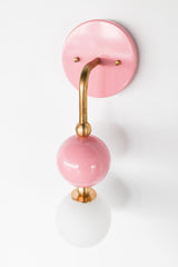 The Beaded Sconce in blush pink stacks a hooked brass arm, small brass ball joints, and a glossy pink powdercoated sphere above a soft white opal glass globe, giving the whole fixture a jewelry-like, beaded silhouette on the wall. It works well as a bedside light in place of a table lamp, beside a bathroom mirror, or anywhere a single statement sconce makes more sense than a matching pair.