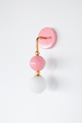 The Beaded Sconce in blush pink stacks a hooked brass arm, small brass ball joints, and a glossy pink powdercoated sphere above a soft white opal glass globe, giving the whole fixture a jewelry-like, beaded silhouette on the wall. It works well as a bedside light in place of a table lamp, beside a bathroom mirror, or anywhere a single statement sconce makes more sense than a matching pair.