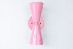 A soft, sweet double-cone wall sconce for a nursery, girl's bedroom, romantic powder room, or mid-century modern space that wants gentle color and crisp, two-directional light. The Dryades sconce pairs an upward and a downward blush-pink cone at a narrow waist, mounted on a round blush backplate with a brass screw accent. Seen from an angle, its bowtie silhouette reads pretty but sharp — a charming pick for grandmillennial, romantic, and playful spaces.