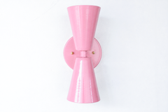 Pink is the easiest way to give a room personality without going loud, and this Blush Pink Dryades sconce does it in a crisp mid-century silhouette. The bowtie shape reads clean and modern, while the soft pink powder coat and warm brass hardware keep it sweet and a little romantic. It's a flexible choice: grown-up enough for a powder room or bedroom, playful enough for a girl's room or nursery. The two opposing cones light up and down, so a pair flanking a mirror or bed brings both symmetry and glow. A char