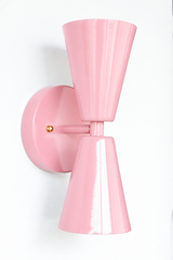 A statement wall sconce for rooms that lead with soft, sweet color and sculptural shapes. This angle of the Dryades sconce shows off its blush-pink double-cone silhouette — one cone throwing light up, one down — meeting at a narrow waist on a round blush backplate with a brass screw accent. Pretty, gentle, and a little retro, it's a charming pick for nurseries, girls' rooms, romantic bedrooms, and mid-century or grandmillennial interiors.
