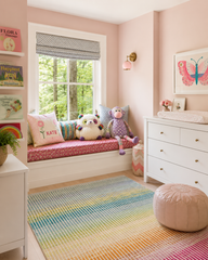 Soft, light-filled nursery reading corner featuring a blush pink Loa sconce with warm brass hardware mounted on a pale pink wall above a gray glider chair. Neutral floor-length drapery, a textured cream rug, blush leather pouf, white crib, and a small round wood side table with pink flowers create a cozy baby girl nursery with modern, feminine, and softly boho style. This pastel nursery design highlights a blush pink wall sconce, colorful nursery lighting, and a stylish reading nook, making it ideal for mod