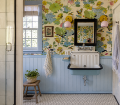 Whimsical powder room with pastel blue beadboard wainscoting, vintage wall-mounted sink, and black-and-white hex tile floor. Two blush pink Loa sconces with warm brass hardware flank a dark framed mirror, adding soft, flattering vanity lighting. Colorful botanical wallpaper with birds and oversized florals brings playful cottage style, grandmillennial charm, and bold pattern mixing to this small bathroom design.