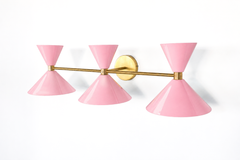 There's something quietly confident about a wide pink vanity light, and this Triple Clancy delivers it in a crisp Italian-inspired silhouette. Seen from the side, the three double-cone shades fan out along a brass bar, each glossy blush pink cone catching the light. It's built for a generous wall, above a long mirror, a double vanity, or a statement powder room, where its scale and color can really register. The brass keeps it warm and timeless, while the pink brings the personality. A great fit for a home 