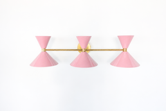 Pink is the easiest way to give a bathroom real personality, and this Triple Clancy sconce makes a statement without losing its mid-century polish. Three glossy blush pink double-cone shades march along a slim brass bar, throwing light both up and down for a soft, flattering glow. The horizontal layout makes it a natural above a wide mirror or a double vanity, and the brass-and-pink combination reads sweet but grown-up. A charming choice for a powder room, primary bath, or any colorful, feminine-leaning spa