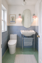 Bright bathroom with blush pink Loa sconces in chrome on either side of an organic wavy mirror above a white sink. Soft pink light spills across crisp white walls, vertical blue tile wainscoting, and a green penny tile floor, tying blush accents to cool-toned, modern bathroom finishes.