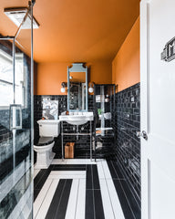 A bold, gallery-like bathroom with dramatic contrasts and confident design choices. Walls are wrapped in black subway tile from floor to ceiling, while the ceiling glows a warm burnt orange that intensifies the space's moody sophistication. Black and white striped floor tiles create strong directional movement down the narrow galley-style room. White pedestal sink with chrome fixtures sits atop black tile, flanked by articulating sconces in chrome with white glass shades that provide task lighting. Dark med