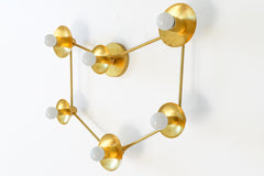 Six-light brass constellation sconce seen from the side, so you can see how each white globe bulb sits out from its round brass cup and how the thin rods hold the points apart. The raw brass has warm patina spots. Use it as a large wall sconce over a bed, sofa or console, or as a flush mount ceiling light. On a white wall it suits mid century modern and space age interiors, celestial bedroom lighting, and eclectic rooms with brass and gold accents.