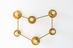 Six-light brass wall sconce shaped like a constellation, with round brass cups and white globe bulbs linked by thin brass rods. The brass is raw and shows warm patina spots, so it reads handmade rather than polished. Mount it on a wall over a bed or sofa, or on the ceiling as a flush mount light. On a plain white wall it fits mid century modern and space age rooms, a celestial or zodiac themed bedroom, and eclectic spaces with warm metal accents.