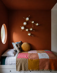 Brass Gemini One wall sconce shaped after the Gemini constellation, mounted above a bed in a color-drenched orange and white cube-style kids room. Warm brass discs and white globe bulbs echo star points, while the handmade patchwork quilt, plaid pillow, cactus pillow, round window, and built-in storage bed add playful maximalist charm. A strong fit for colorful kids room lighting, zodiac-inspired wall lights, brass constellation sconces, eclectic nursery design, modern playroom decor, and artful statement w