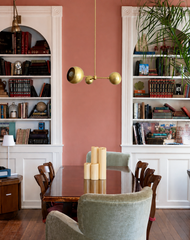 This dining room reads like a well-used library, with two dense, symmetrical bookcases flanking a deep terracotta pink accent wall. The Loa Chandelier hangs at the center in solid brass, its asymmetrical orb shades adding movement without competing with all those books. Mismatched dining chairs, velvet armchairs, and a stack of pillar candles keep the room feeling collected and lived in rather than staged for a photo.
