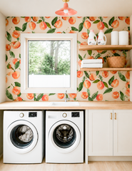 A laundry room wrapped in peach and cream fruit-print wallpaper, with a farmhouse-style window over the sink, open wood shelving, and white cabinetry with brass pulls. Overhead, a peach flushmount with brass hardware picks up the wallpaper's warm tone instead of defaulting to a plain white fixture. Proof that a laundry room can be just as pretty as the rest of the house.
