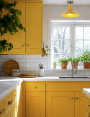 A butter yellow shaker kitchen with white subway tile, white countertops, and a sink window lined with potted herbs. Overhead, a yellow flushmount with chrome hardware picks up the exact shade of the cabinetry, tying the whole room together instead of defaulting to a neutral fixture. A cheerful, fully committed color kitchen for anyone tired of white and gray.